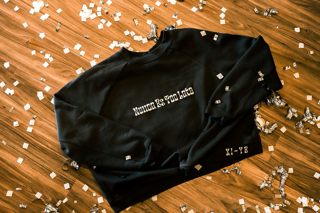 Grey crewneck on the floor with confetti with the saying " Nuncio Es Too Late" on the front. 