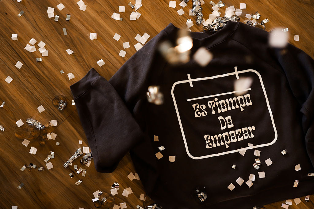 Grey crewneck on the floor with confetti with the saying " Es Tiempo De Empezar". 