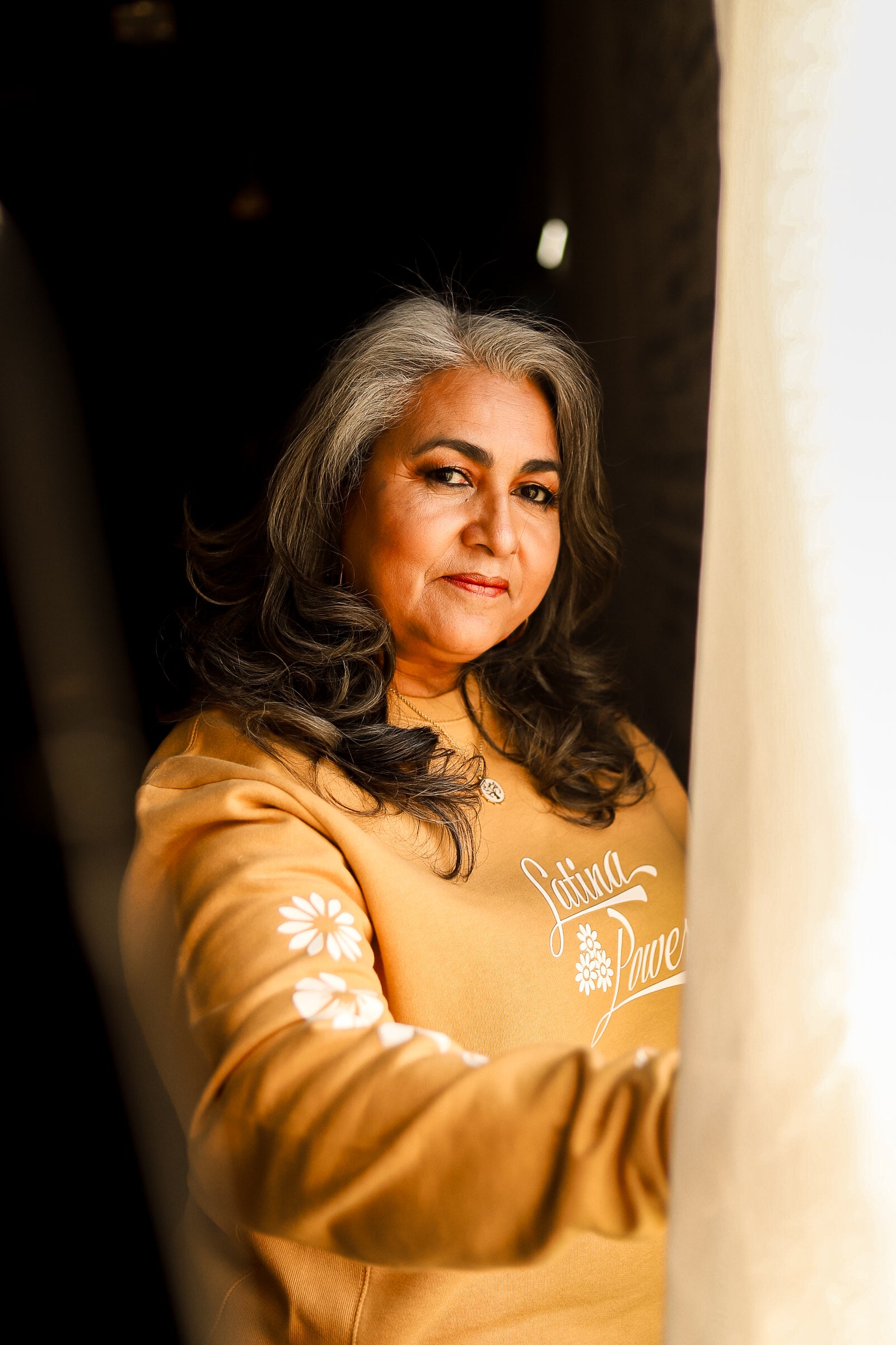 Person leaning toward a window with camel yellow crewneck with white flowers and "Latina Power' saying. 